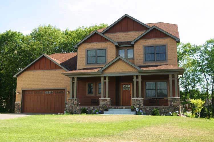 Two-story brown house with attached garage and front porch, set on a green lawn with trees behind.