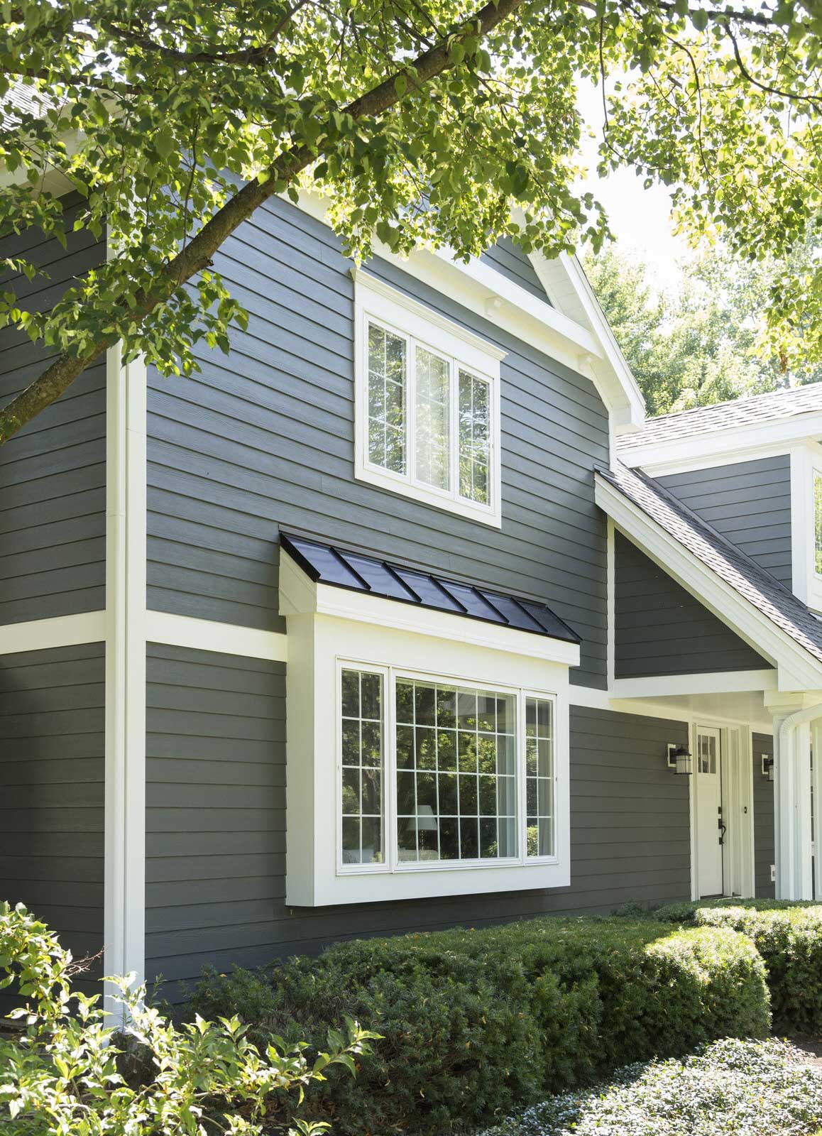 Gray house exterior with white trim, front window, and green shrubs under leafy trees