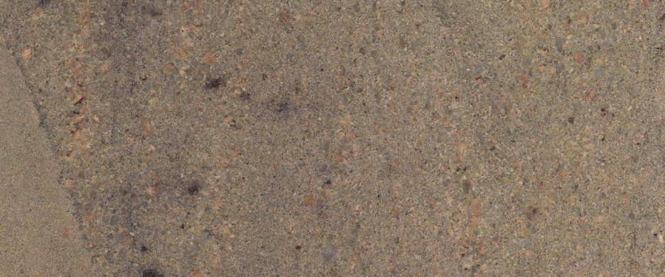 A close-up view of textured, sandy-beige stone or concrete surface with fine, mottled dark and light grains.