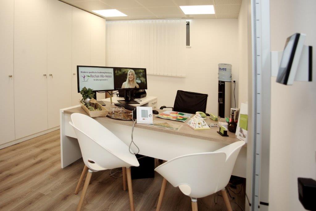 Beyaz mobilyalı, masalı ve iki sandalyeli ofis. Bilgisayar ekranında bir kadın görülüyor.