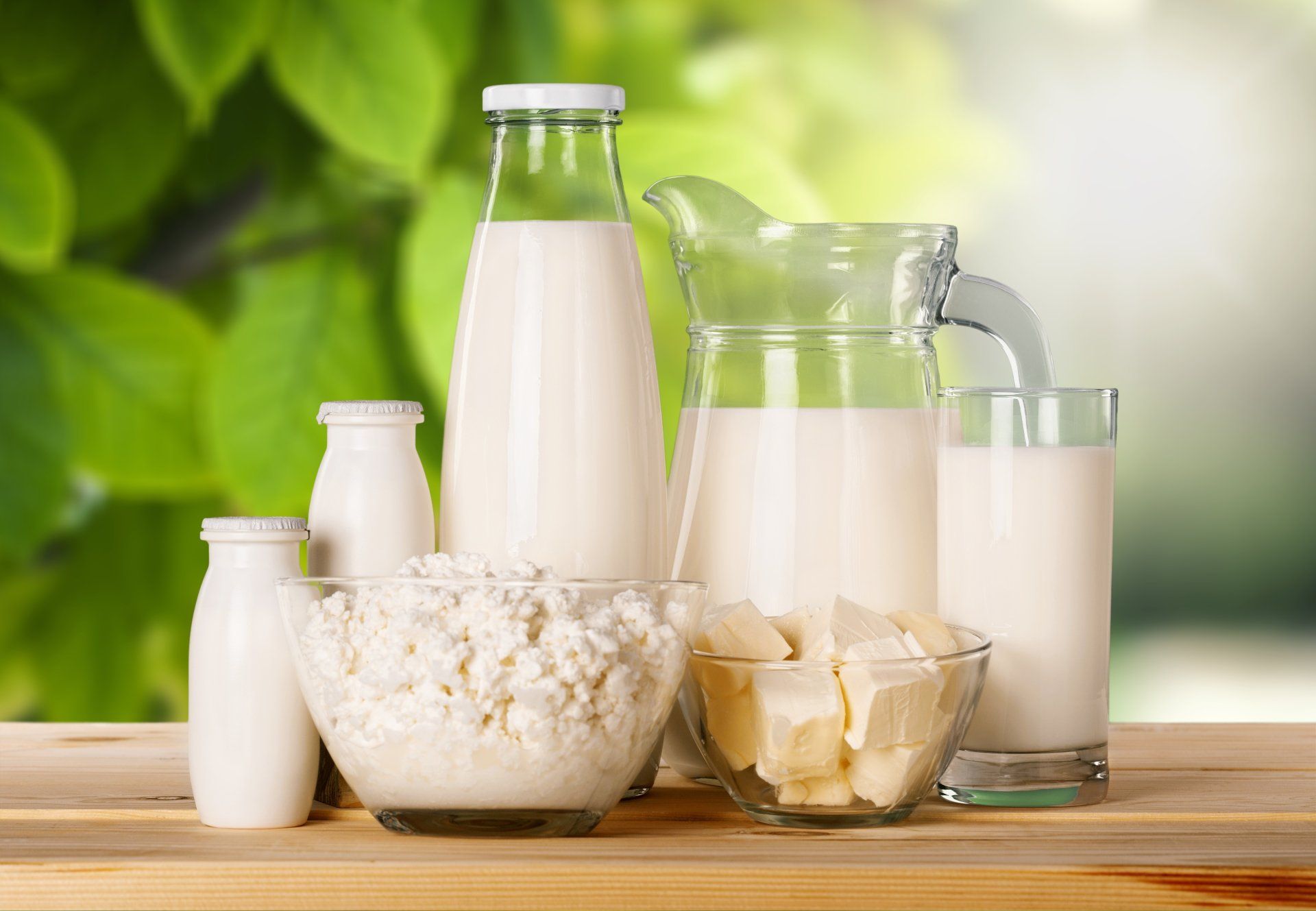 Разложенные на деревянной поверхности молочные продукты, включая бутылки с молоком, кувшин, стаканы, творог и масло.