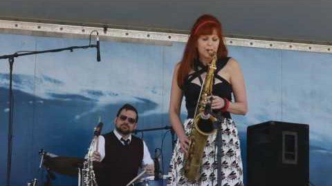 A woman is playing a saxophone on stage while a man plays drums.