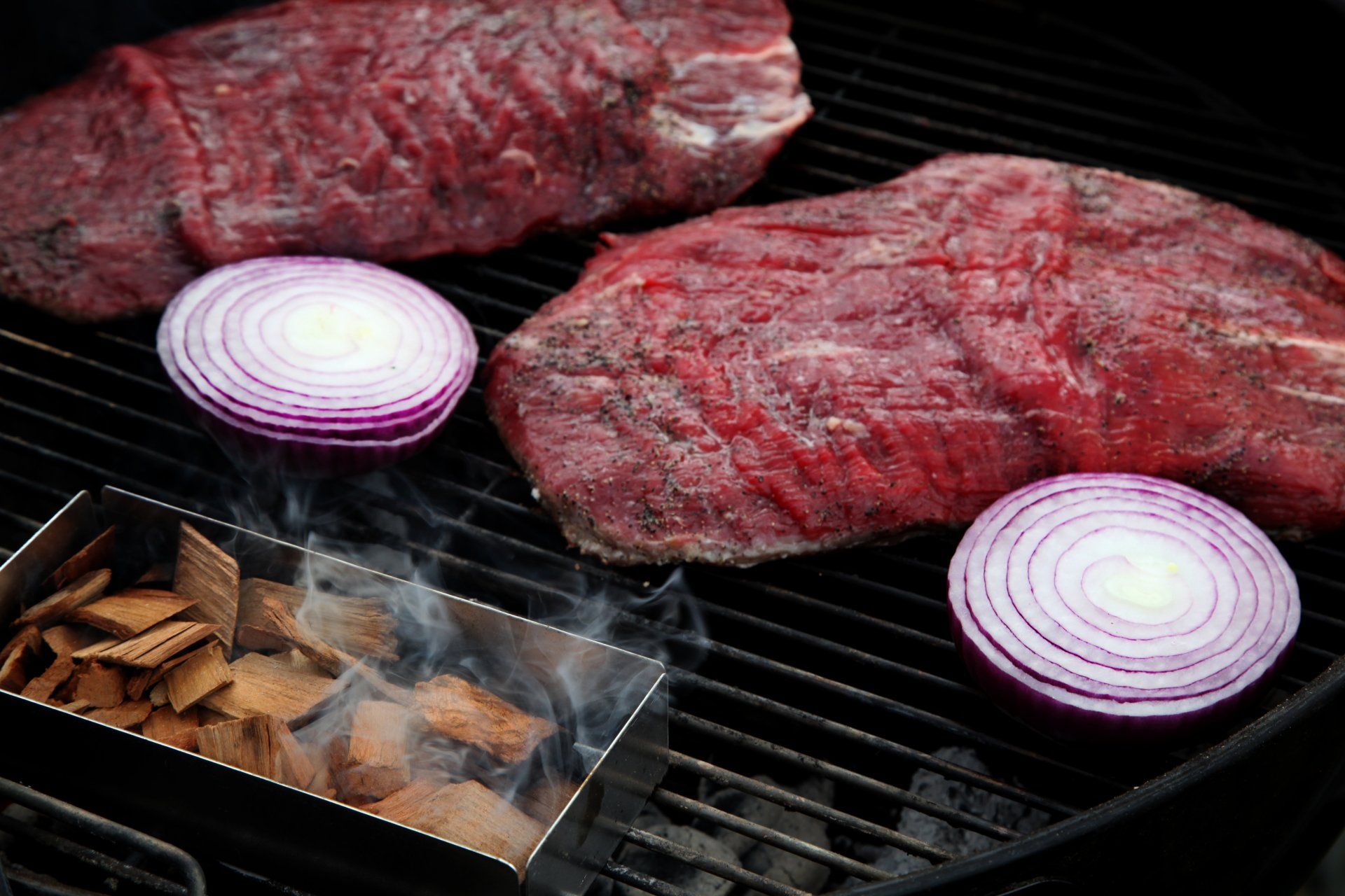 Closeup of a smoking tray full of smoking chips and two flank steaks and two red onions on the grill