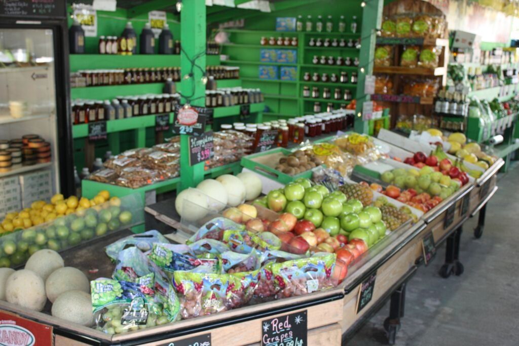 Produce stand with various fruits and vegetables, green shelving, and assorted jars in the background.
