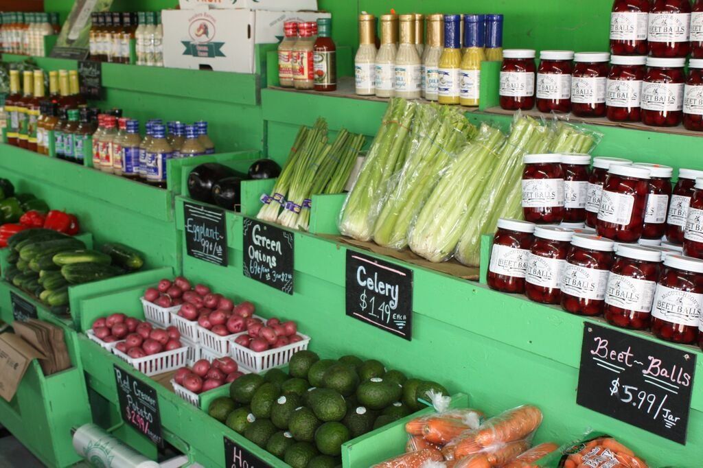 Green produce stand displaying vegetables, jars of preserves, and sauces.