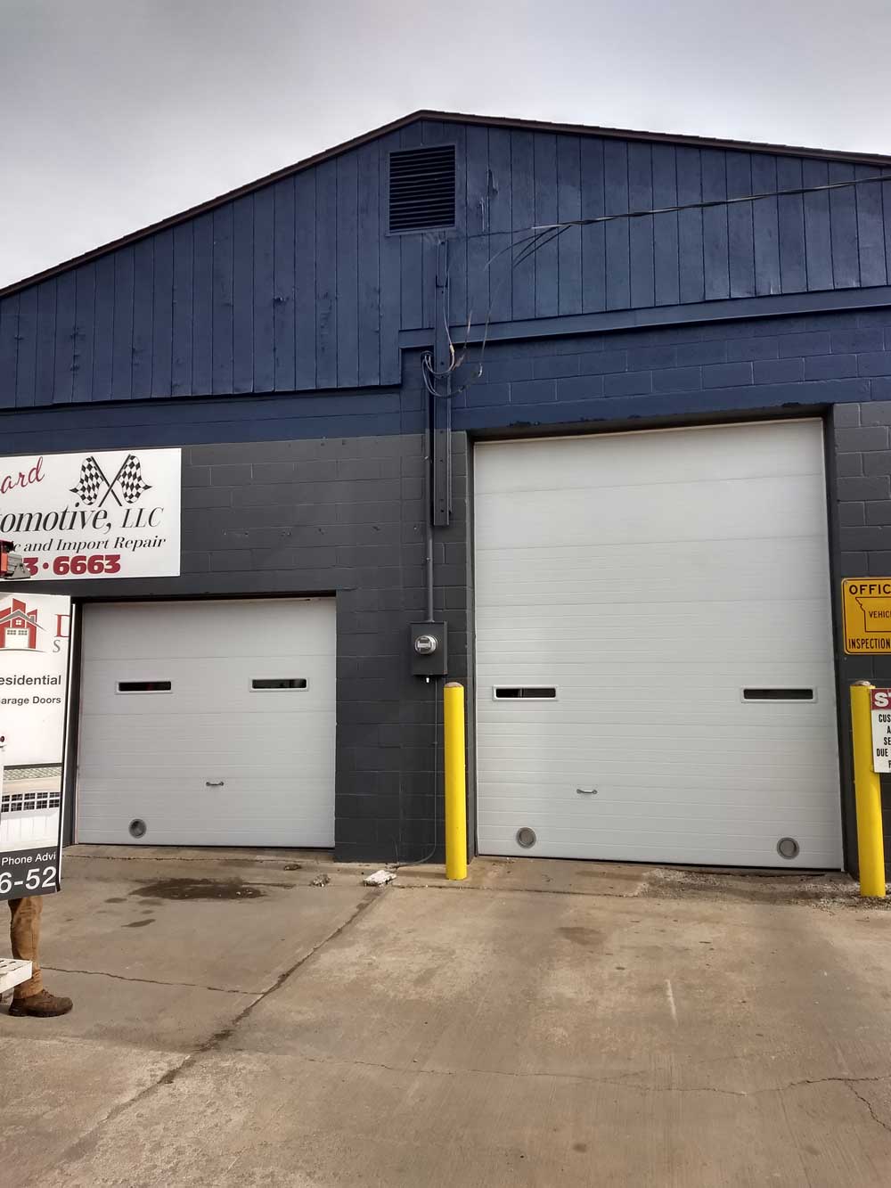 Industrial building with two gray garage doors and a blue metal facade, viewed from a concrete lot