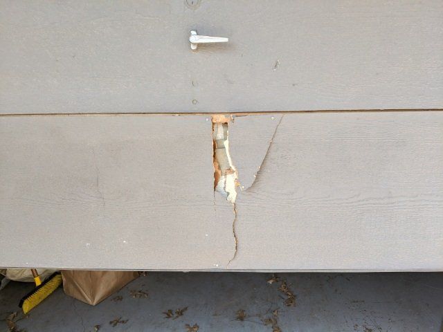 Damaged gray siding on a house with a vertical crack and exposed wood near a small pipe fitting