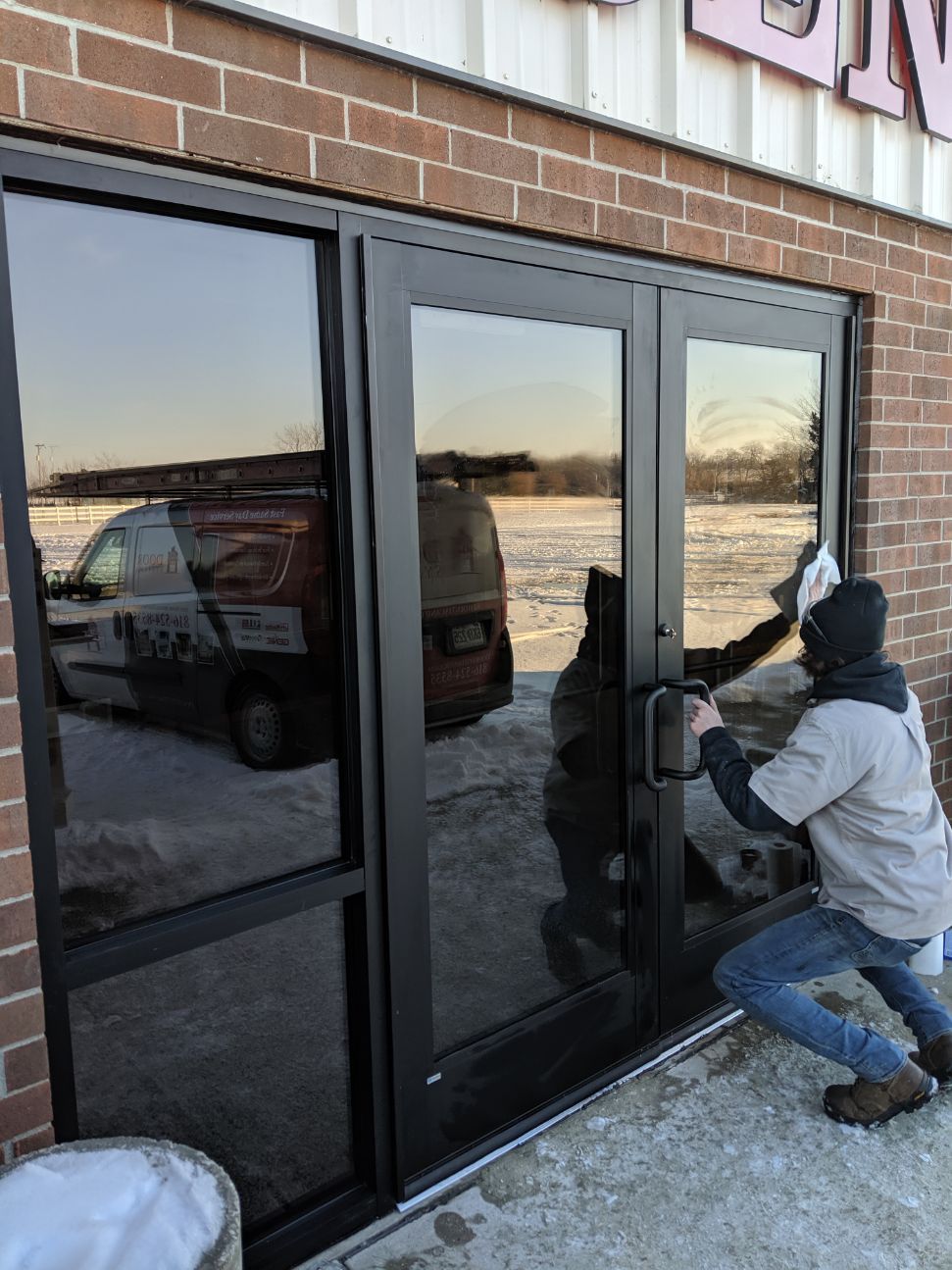 Person kneels at a glass storefront door, prying at the handle with a tool in a snowy parking lot.