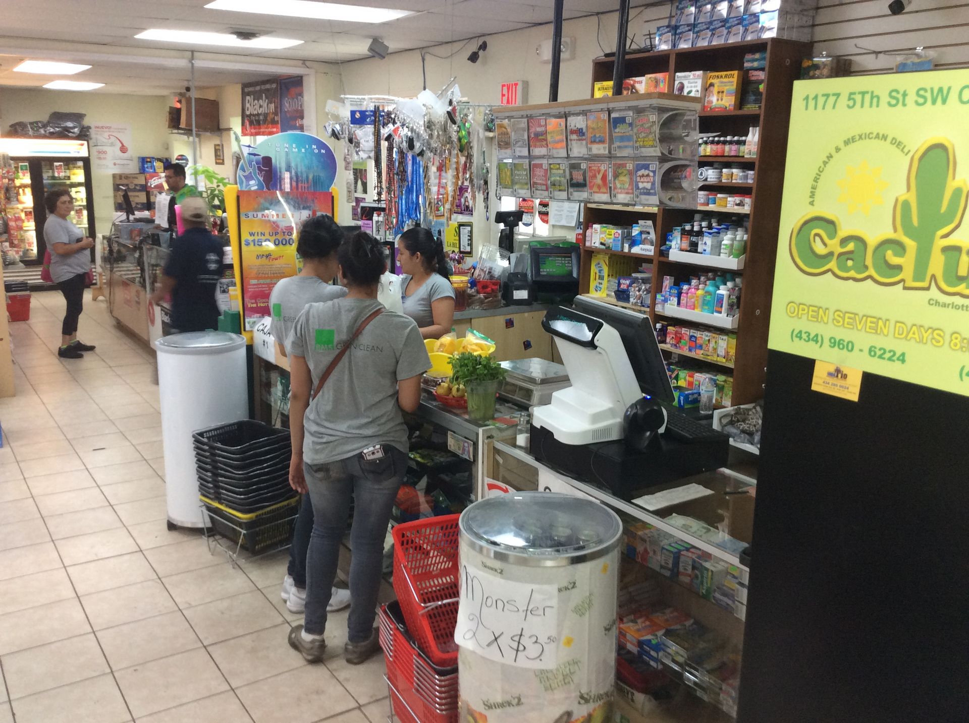 Inside a small store with customers at a counter. Display shelves filled with products, a refrigerator, and a Cactus sign.