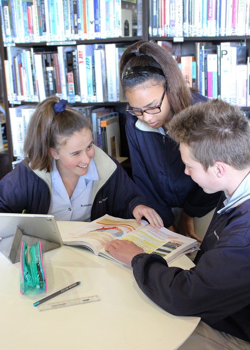 three students are sitting at a table in a library looking at a book .