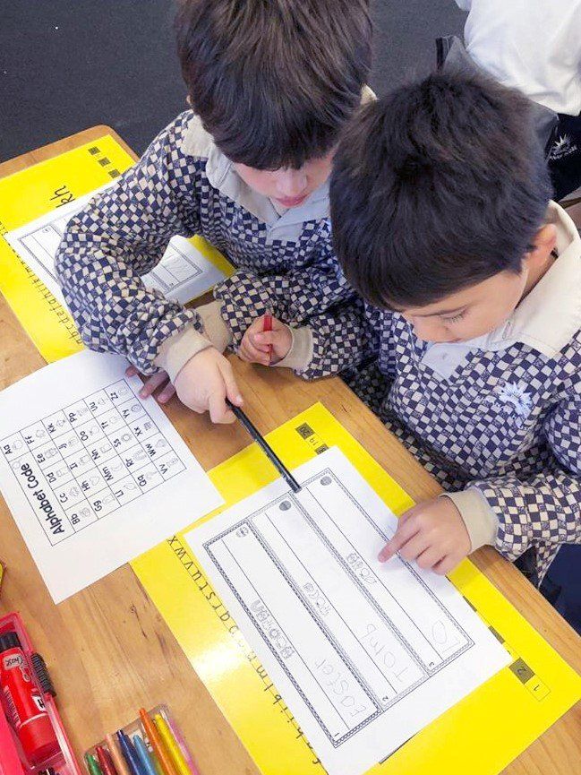 two young boys are sitting at a desk writing on a piece of paper