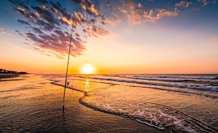 Sunset over the beach with a fishing pole. Orange and gold sky reflecting on wet sand and waves.