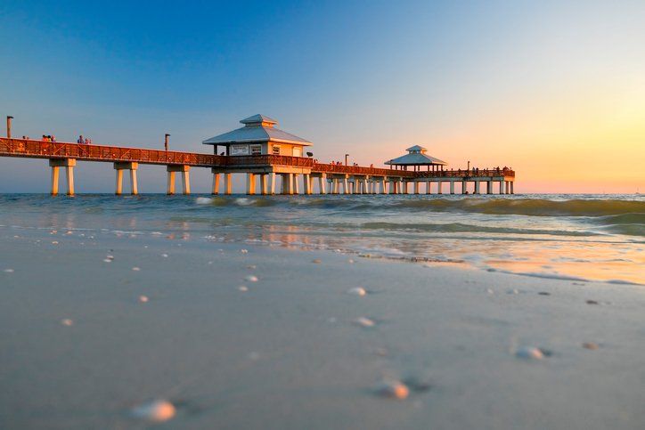 Beach scene at sunset: a long pier with two gazebos extends into the ocean, people silhouetted on it.