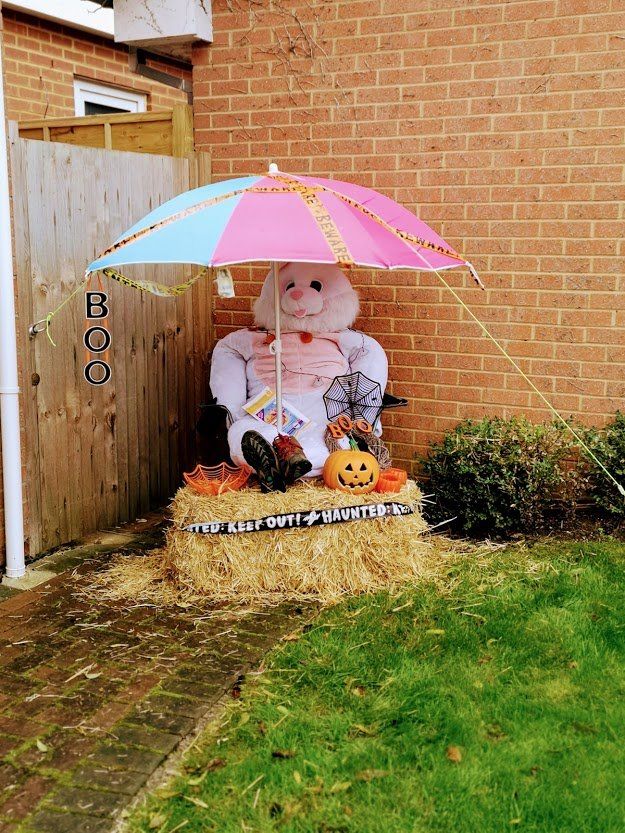 A bunny scarecrow under a pink and blue umbrella, on a hay bale, with a pumpkin and spiderwebs.