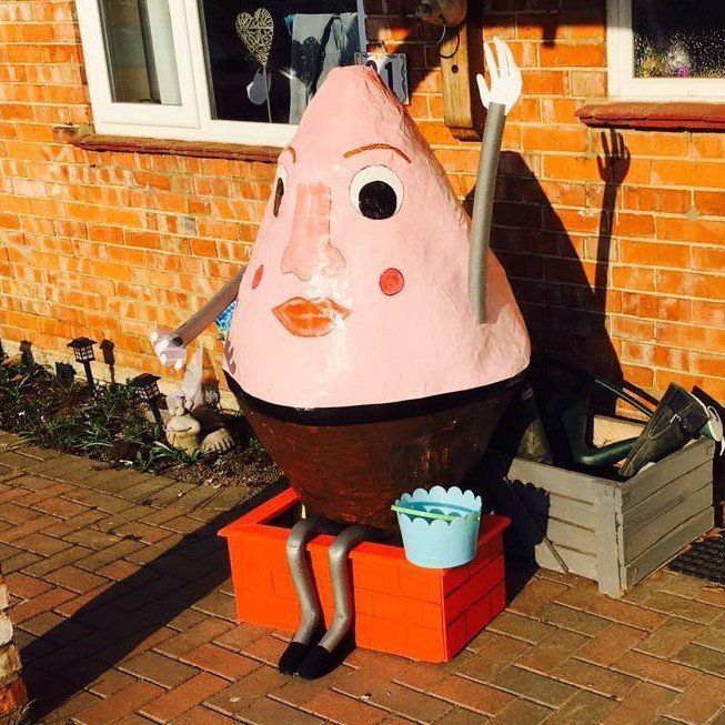 A whimsical, pink-headed cone-shaped figure with a large nose and arms raised, seated on a red box.