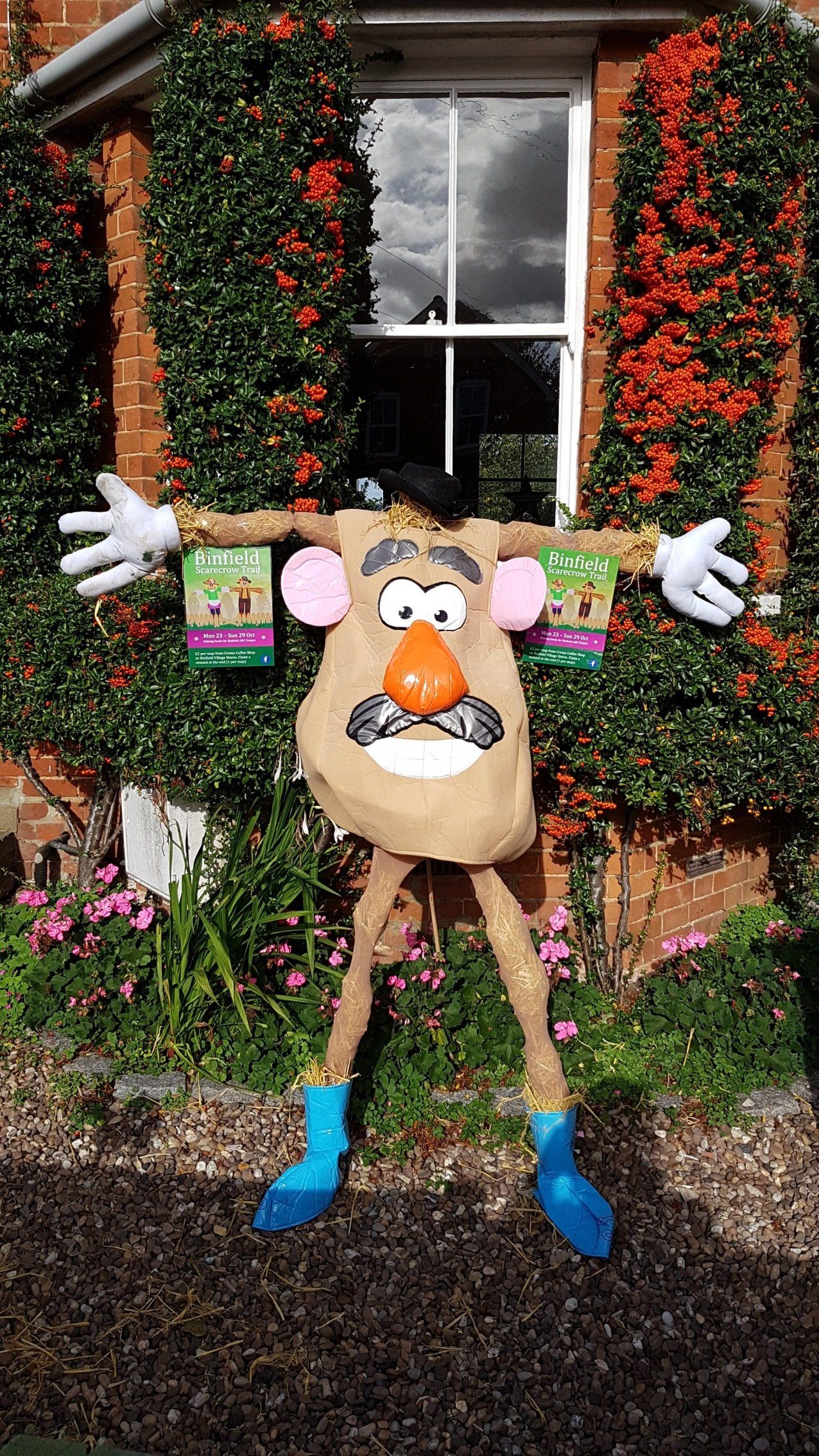 Mr. Potato Head scarecrow with arms outstretched, standing in front of a window and brick building covered in greenery.