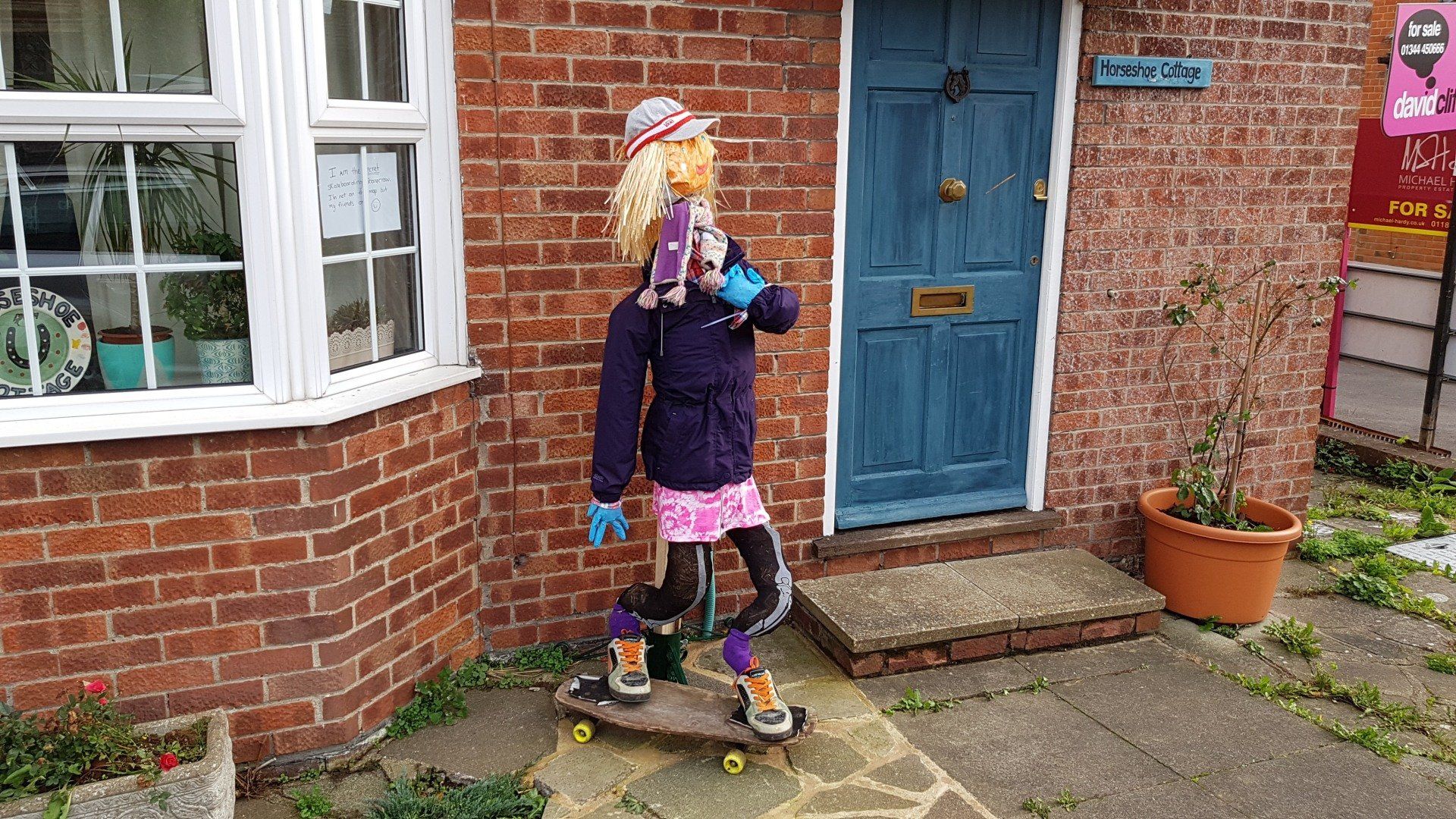 Scarecrow on a skateboard in front of a brick building with a blue door and a white window.