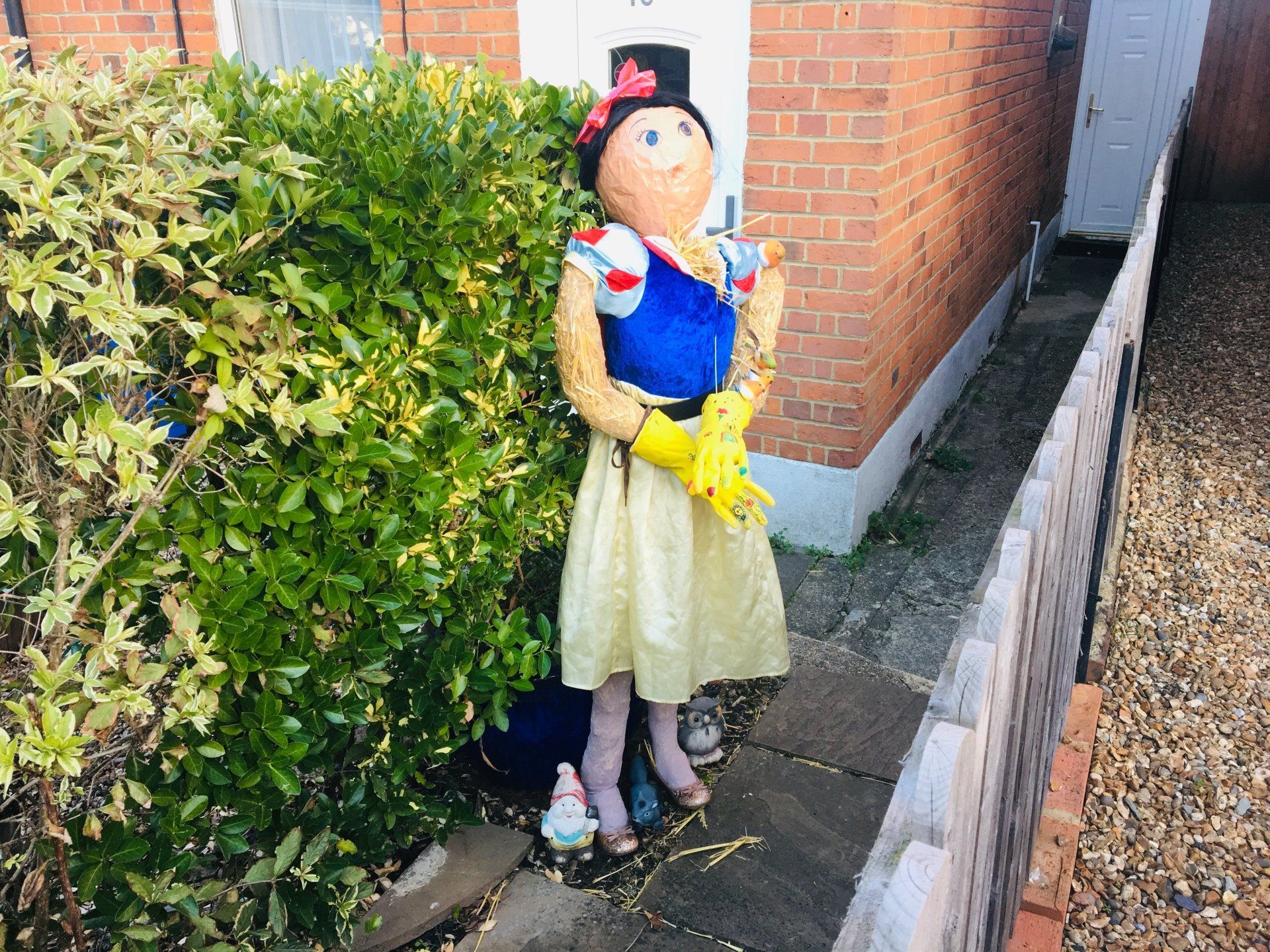 Snow White scarecrow with a top hat, yellow gloves, standing beside a bush and a house.