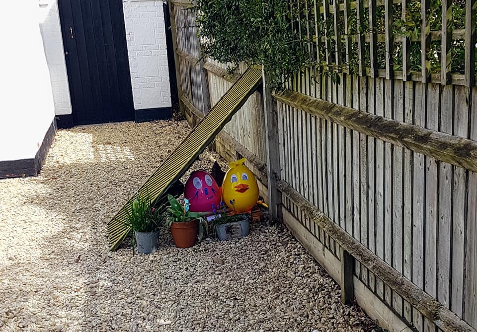 Easter eggs and plants next to a wooden fence and doorway in a gravel yard.