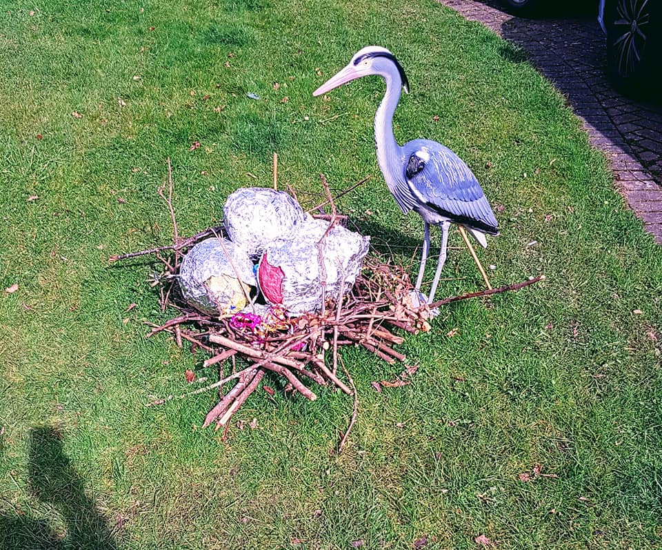A garden display of a heron statue next to a nest with three decorated eggs, on grass.
