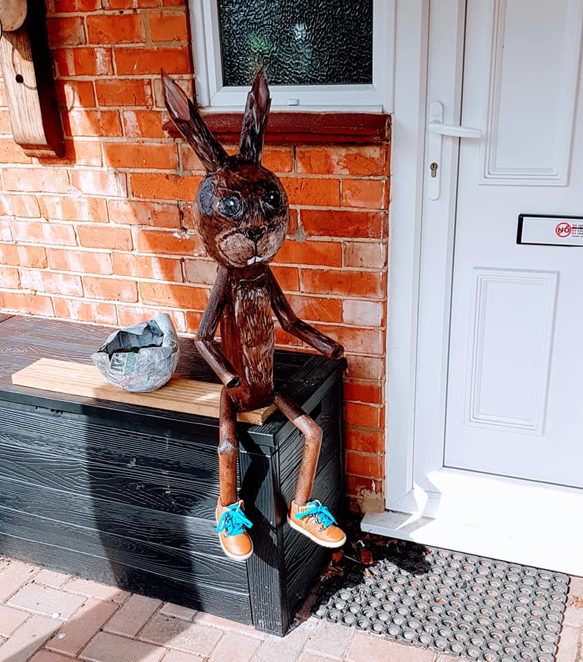 Metal rabbit sculpture wearing blue and tan sneakers sits on a black box near a brick building.
