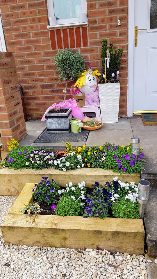 Entryway with flower boxes and a brick wall. Viola flowers in wooden planters, a tiered garden display, and a decorated balloon.