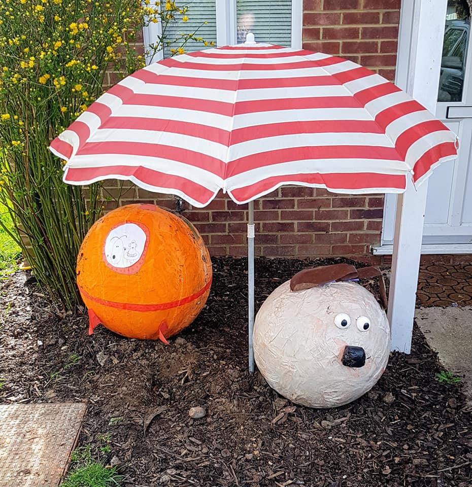 Two round sculptures: an orange one and a sheep, under a striped beach umbrella in a garden.