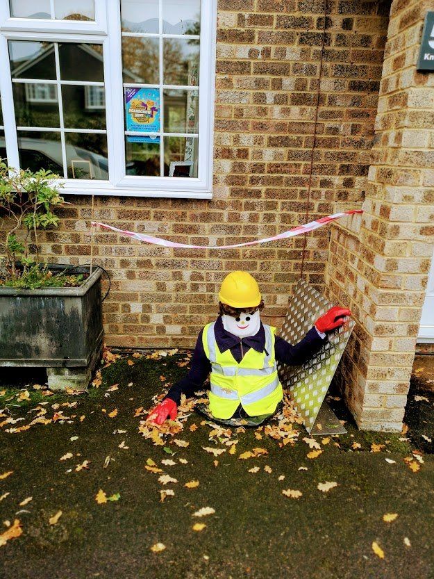 Scarecrow dressed as a construction worker emerging from a hole, yellow helmet and vest, red gloves, with caution tape.