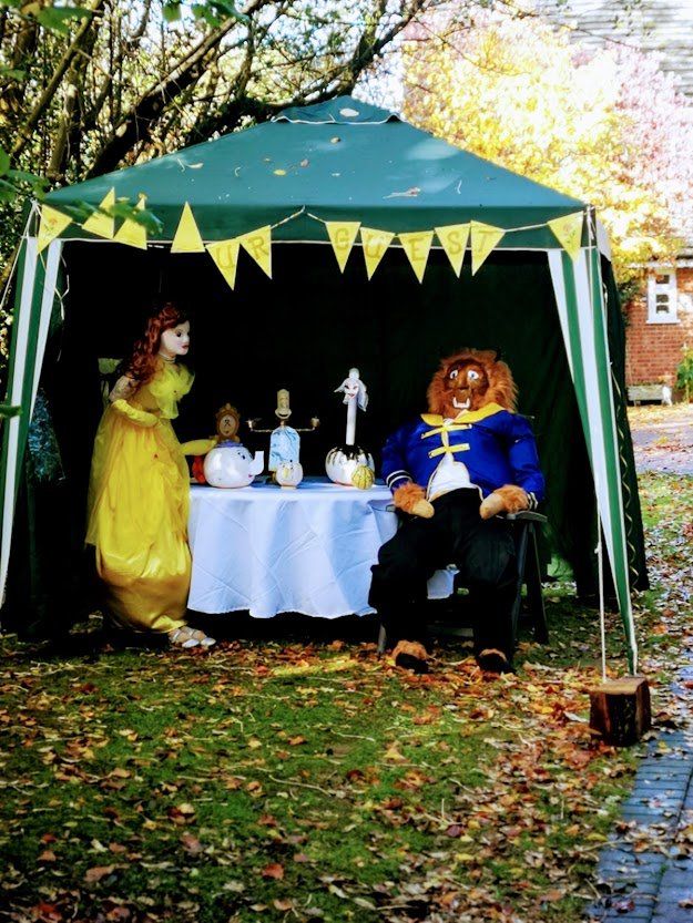 Beauty and the Beast characters in a tent decorated with pennants; Belle looks at Beast sitting, a table with figurines between them.
