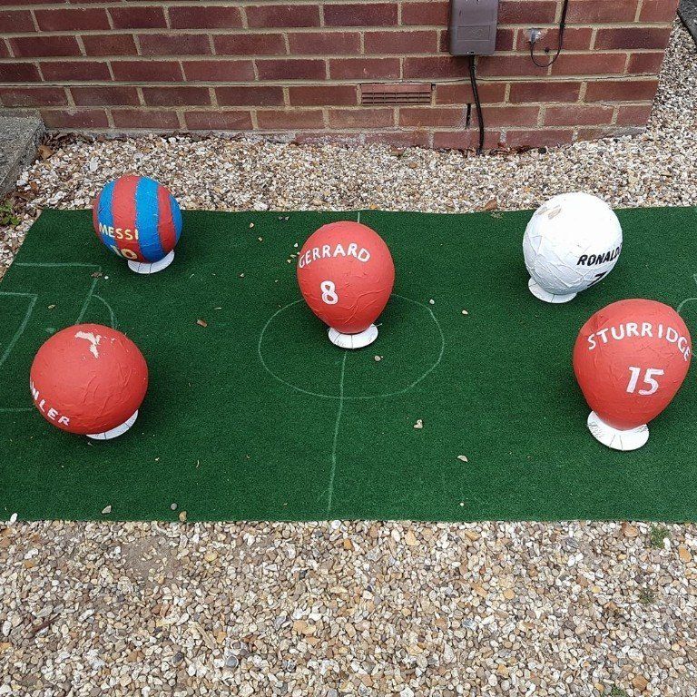 Five soccer ball sculptures on a green field. Four are red, one is blue and red striped.