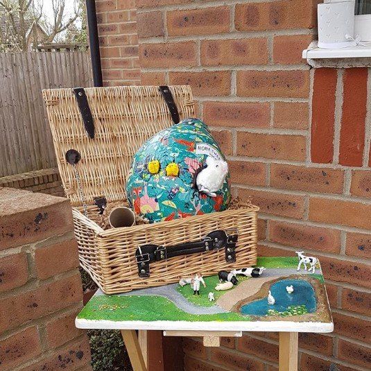 Wicker basket with decorated egg, a small table with a miniature farm scene, outside near a brick wall.