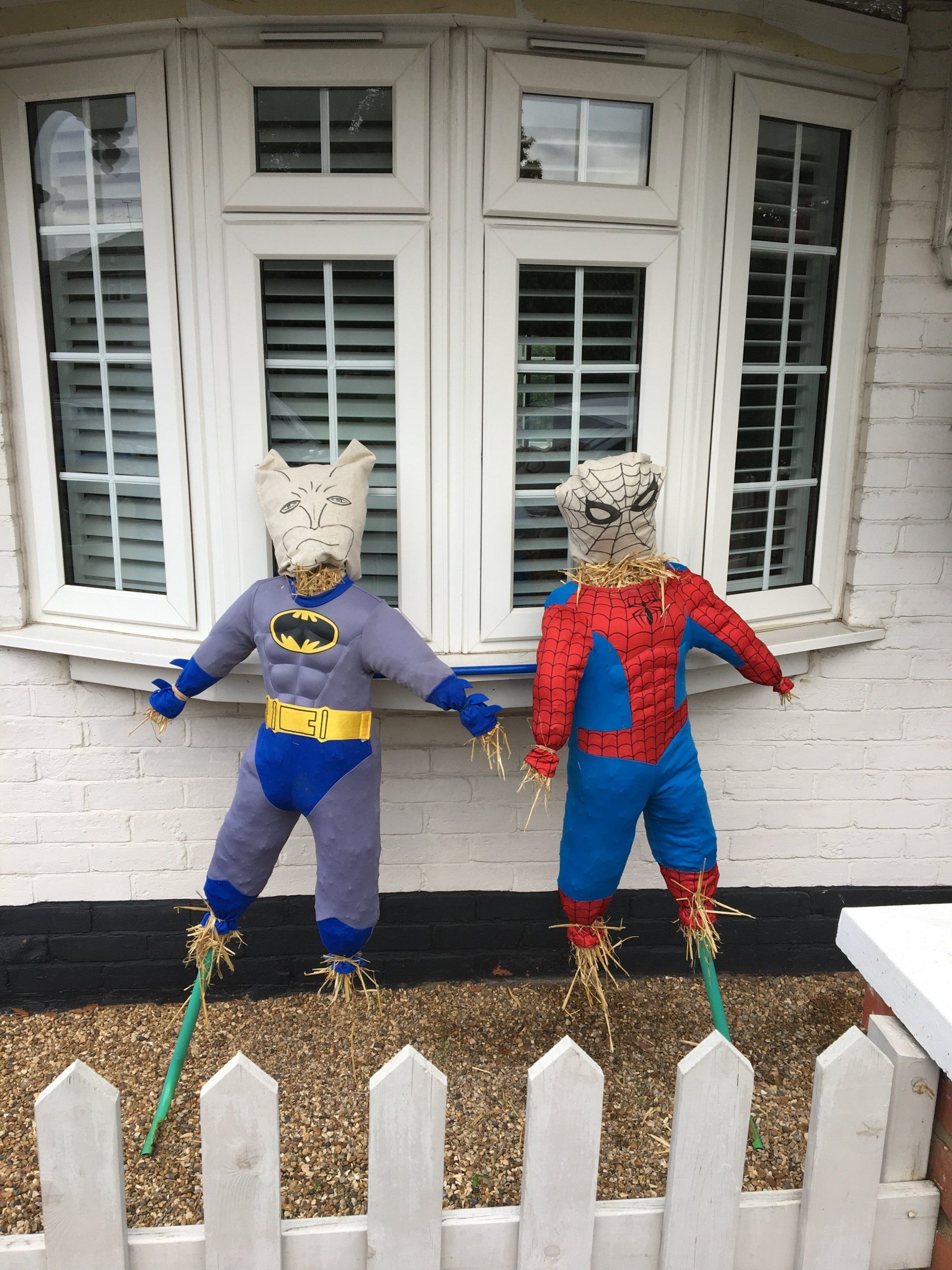 Scarecrows of Batman and Spider-Man stand in front of a white house with a picket fence.