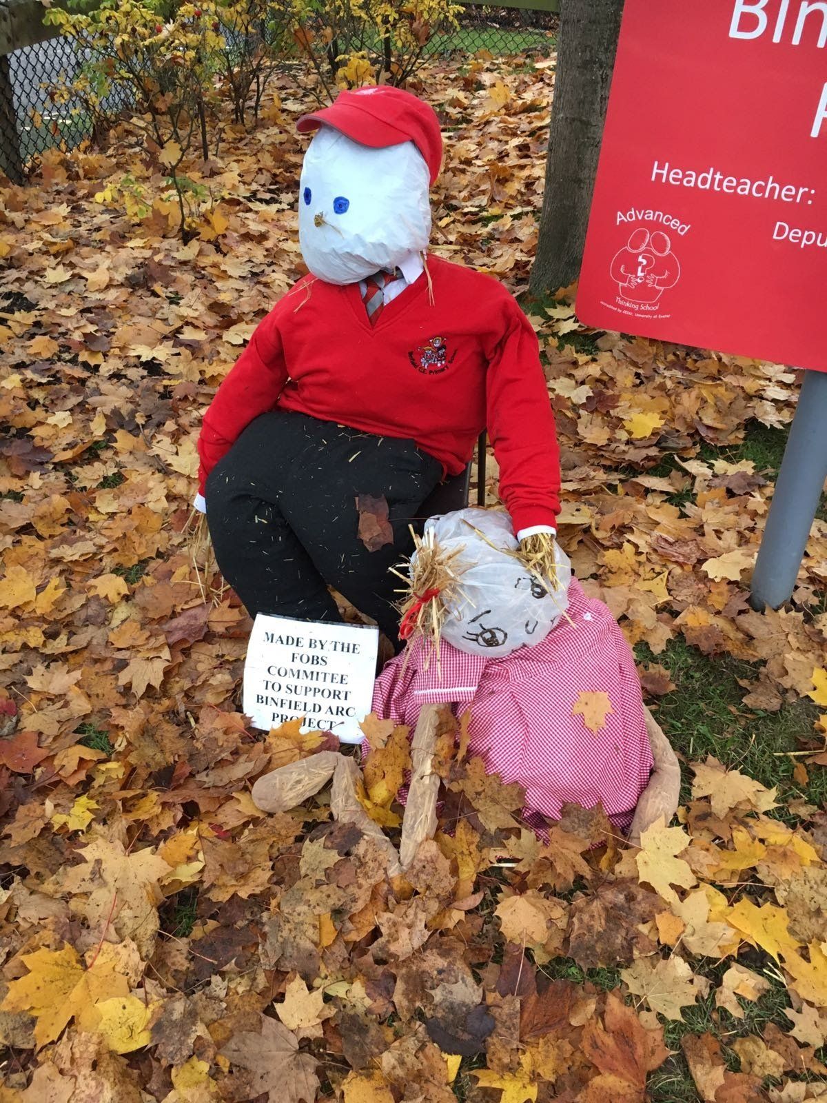 Two scarecrows in a leaf-covered setting; one standing wearing red, and another lying down in pink.