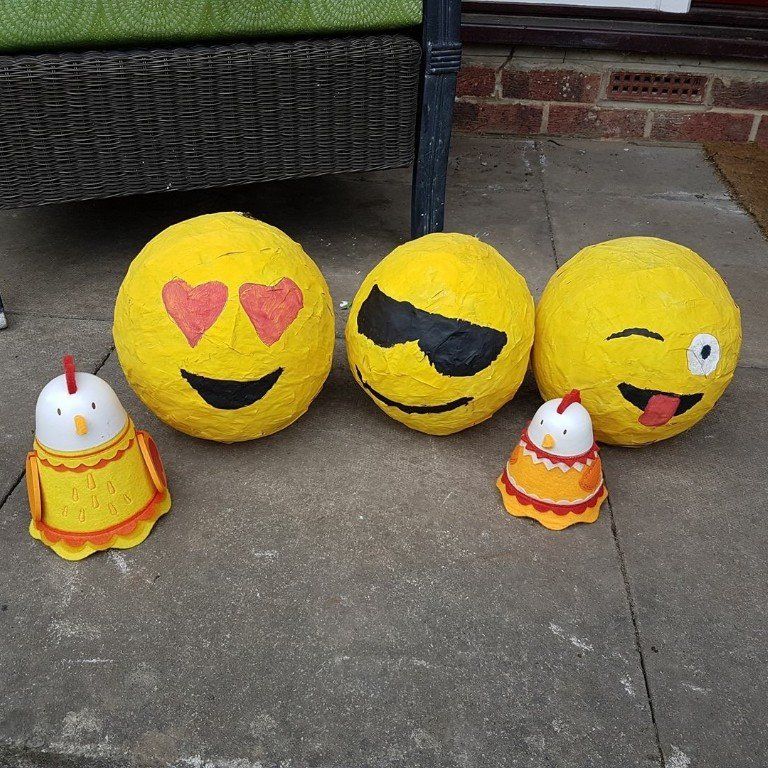 Three yellow emoji balls and two chicken figurines on a patio. The emoji balls have heart eyes, sunglasses, and a wink.