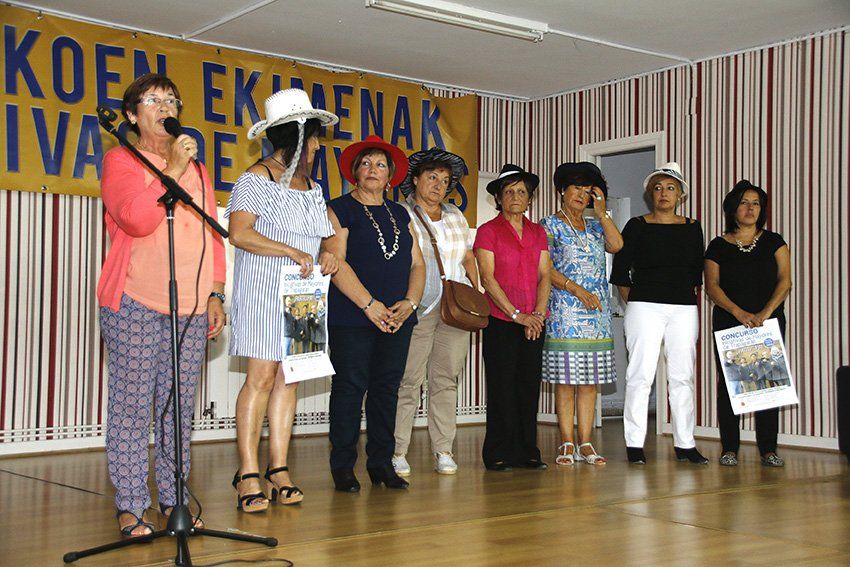 Grupo de mujeres en el escenario con sombreros, una de ellas hablando por un micrófono, frente a una pancarta.