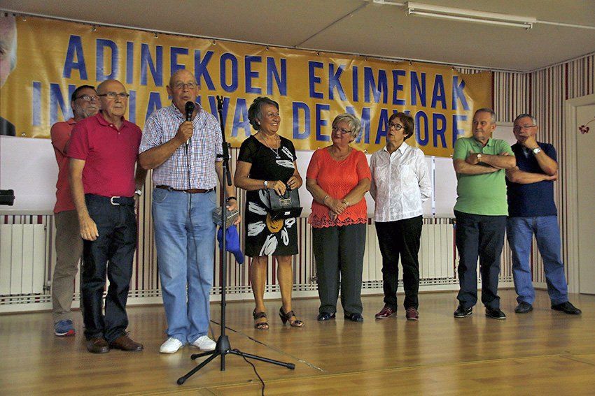 Grupo de personas de pie en el escenario de una habitación con una pancarta y un hombre hablando por el micrófono.