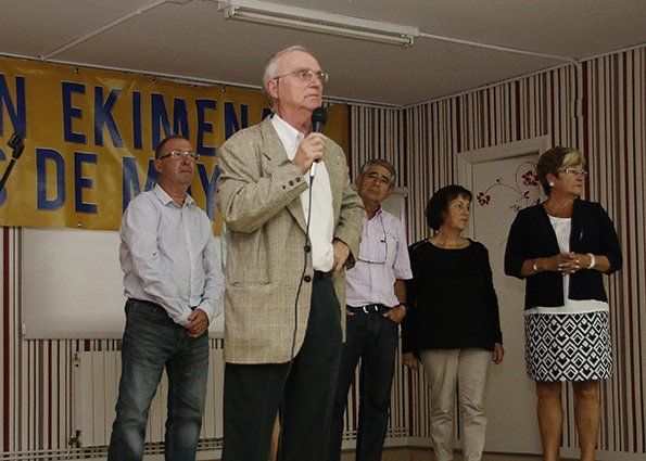 Hombre hablando por un micrófono, rodeado de un grupo de personas en un escenario con una pancarta.