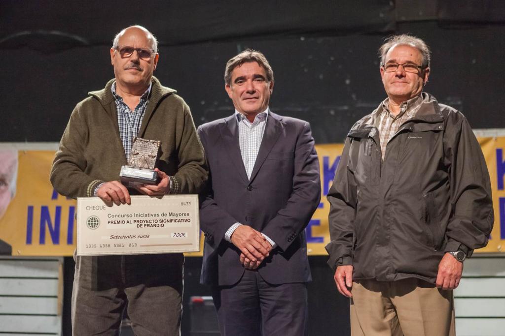 Tres hombres en el escenario sosteniendo un premio y un cheque; uno en el centro, con traje, flanqueado por hombres con chaqueta.