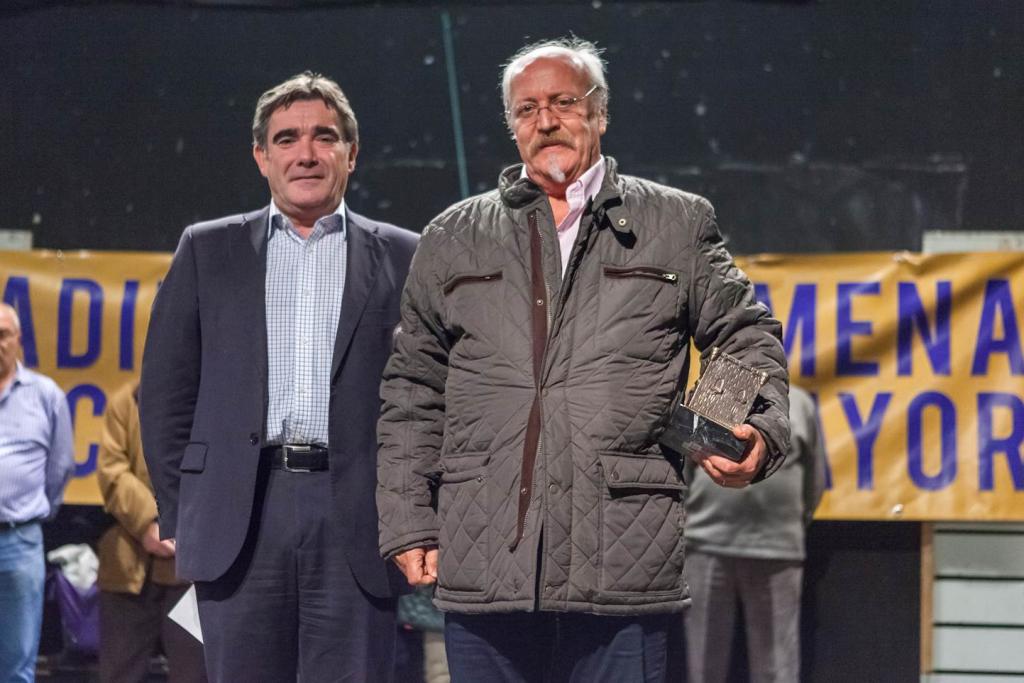Dos hombres de pie, uno de ellos con un trofeo. Traje oscuro y chaqueta acolchada. Fondo con el texto