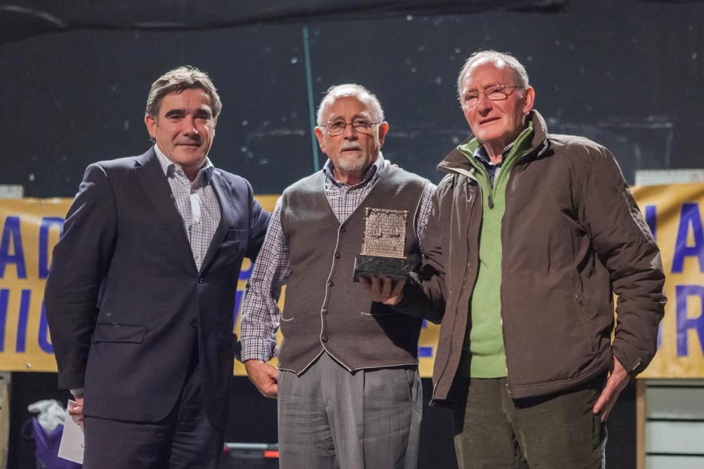 Tres hombres en una ceremonia de premios, uno de ellos con un trofeo. Están de pie frente a una pancarta.
