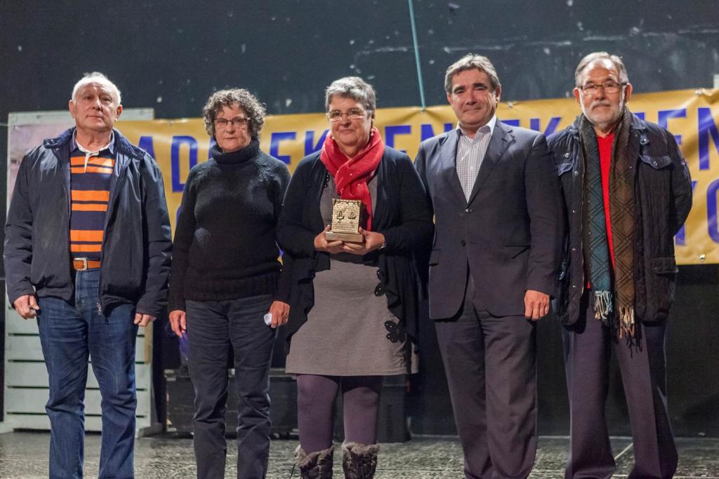 Cinco personas en un escenario, sosteniendo un premio. La mujer del centro lo sostiene. El fondo es oscuro.