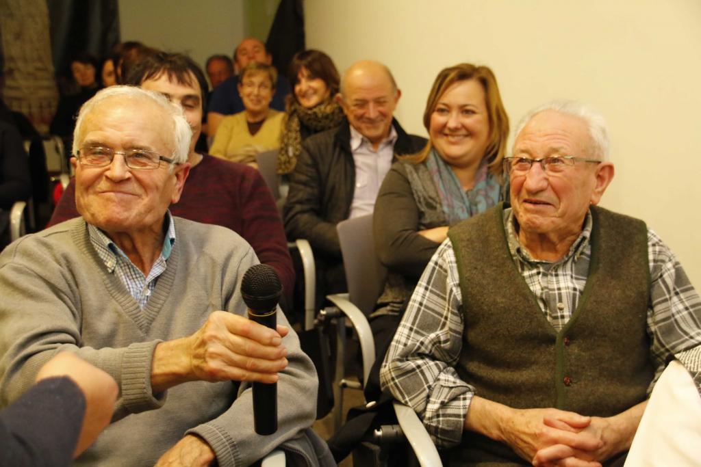 Dos hombres mayores con micrófonos sonriendo en un evento, con otros en el fondo.