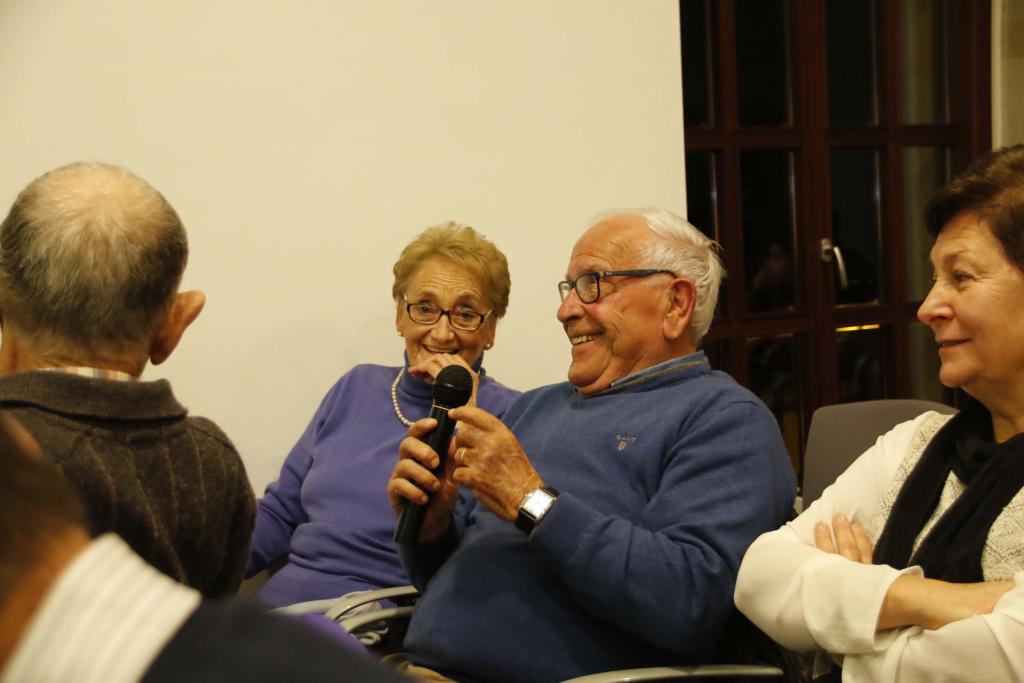 Un anciano habla por un micrófono en un evento, sonriendo. Sentado junto a una mujer y al lado de otra.