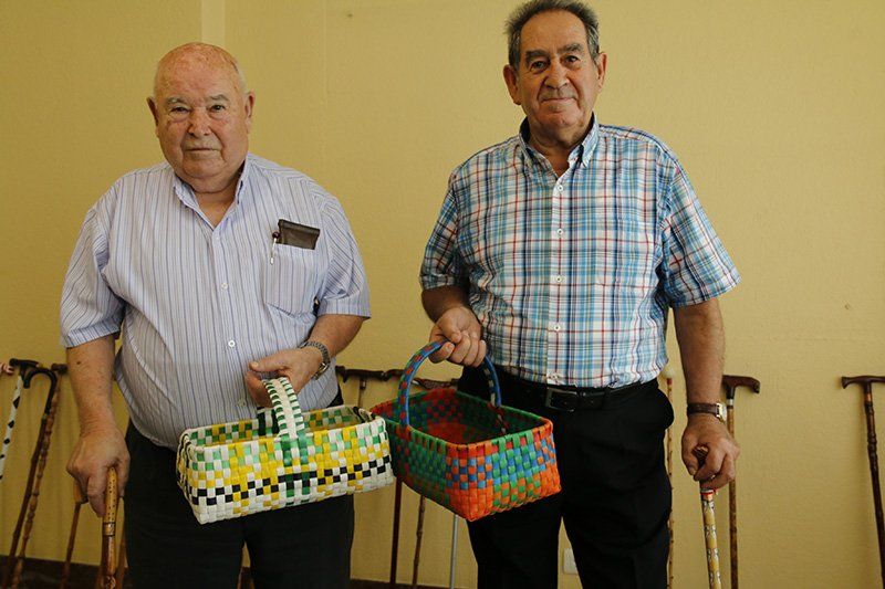 Dos hombres mayores sosteniendo cestas tejidas de colores, de pie con bastones contra una pared.