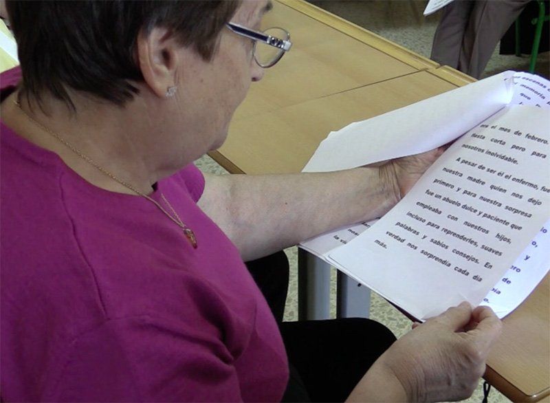 Una mujer con gafas lee documentos, posiblemente sentada en un escritorio. Tiene el pelo corto y lleva una blusa rosa.