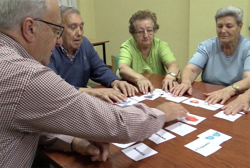 Cuatro personas mayores jugando a las cartas en una mesa, mientras un hombre señala una carta.