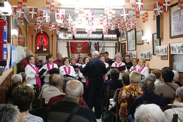 Coro cantando en una sala decorada con público. Vestimenta blanca y rosa. Banderas ondeando en el cielo.