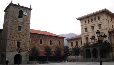 Torre de iglesia de piedra y edificio en una plaza de la ciudad; otro edificio a la derecha, árboles y cielo nublado.