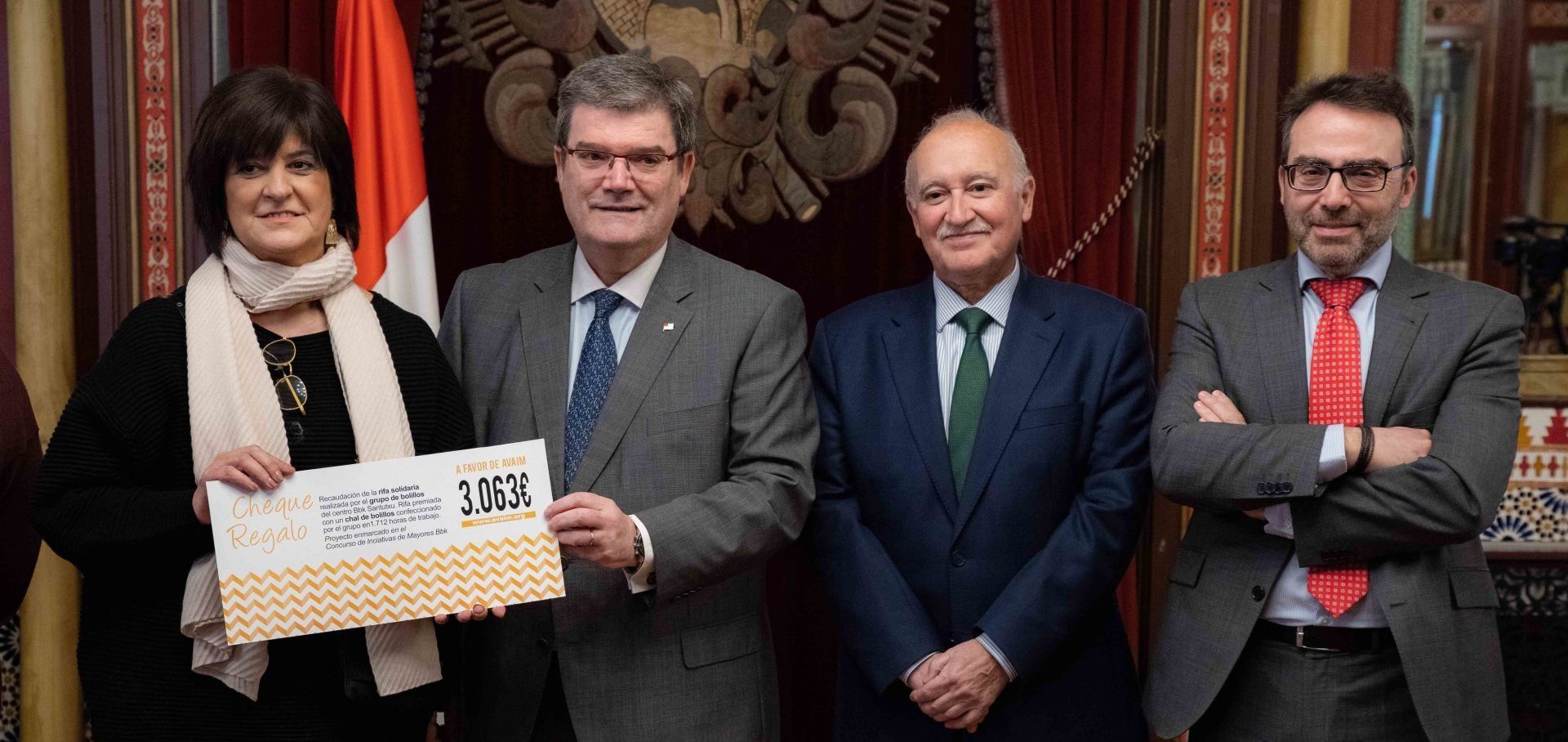Cuatro personas vestidas de traje posan sosteniendo un documento en un entorno formal con un escudo y una bandera.