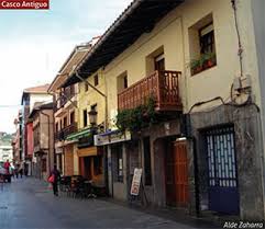 Escena callejera con edificios, tiendas y gente caminando. Los edificios tienen balcones, ventanas y puertas.